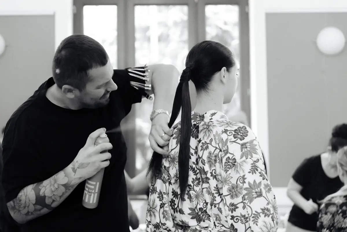 Attila fixează cu spray o coadă lucioasă, ținând agrafele în mână, în culisele unei prezentări