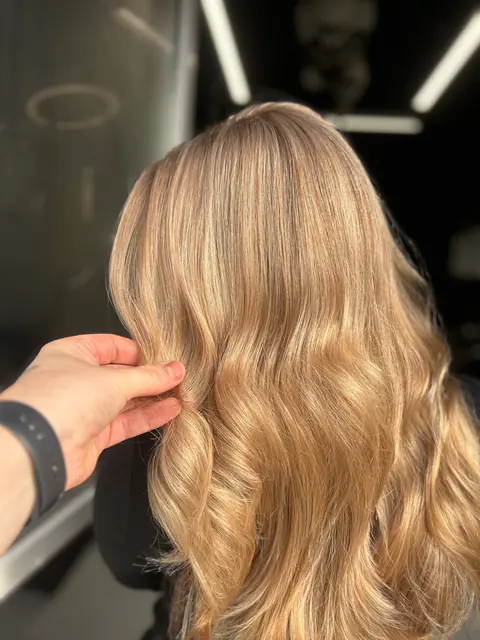 Stilistul ridică o șuviță de păr blond miere, cu valuri aurii, pentru a arăta culoarea