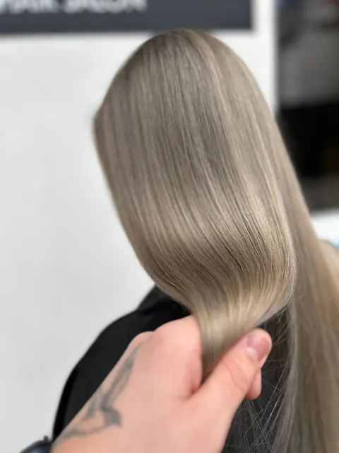 Mână tatuată care ține o șuviță de păr drept, lucios, blond bej-cenușiu (mushroom blonde)