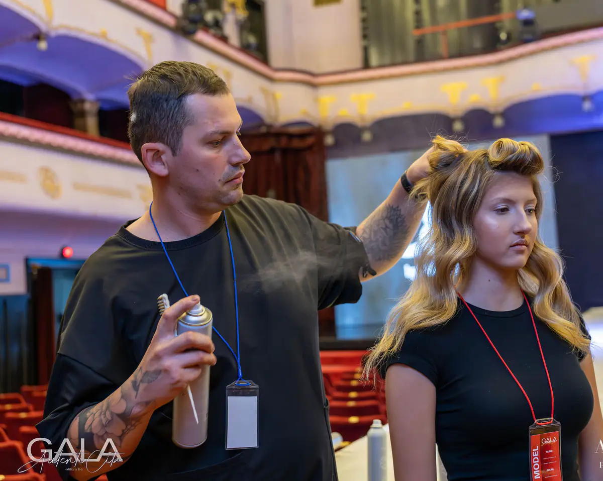 Attila fixează cu spray valurile unui model pe scena unui teatru, în timpul unei gale